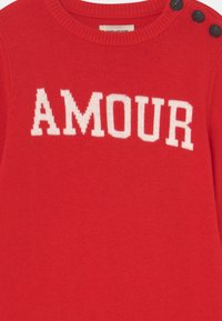 Czerwony sweter z dzianiny z napisem "AMOUR" w białym kolorze. Posiada trzy czarne guziki na lewym ramieniu. Gładka tekstura.