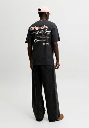Personne portant un t-shirt graphique noir, un jean noir large, des chaussures marron clair et une casquette rose pâle, debout devant un fond blanc uni.