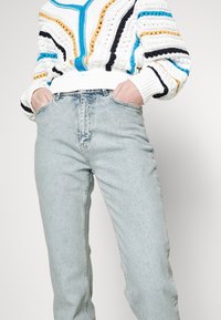 Personne portant un jean taille haute bleu clair avec les mains dans les poches et un pull en tricot blanc à rayures bleues, noires et moutarde.