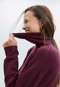 Frau mit langen braunen Haaren, die den Kragen ihres lila Rollkragenpullovers anhebt, lächelt und nach rechts schaut, vor einem klaren Himmel.