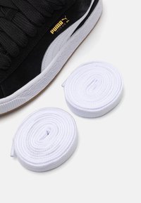 Baskets Puma en suède noir avec de grosses lacets, dotées d'une bande blanche et d'un logo doré. Deux lacets blancs enroulés placés à côté de la chaussure.