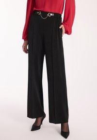 Czarne spodnie o szerokich nogawkach z teksturowanej tkaniny, elastycznym pasem w złotym odcieniu i bocznymi kieszeniami. W zestawie z czarnymi szpilkami.
