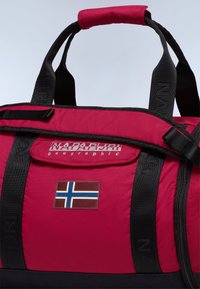 Punainen nylontyylillä varustettu kassi mustilla hihnoilla, jossa on etutasku, jossa on Norjan lipun merkki ja "Napapijri geographic" -logo.