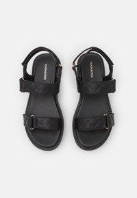 Černé sandály s prošívanými popruhy, dvěma nastavitelnými sponami a hladkou gumovou podrážkou. Navrženy s uzavřenou patou a otevřenou špičkou.