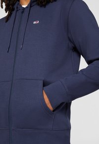 Felpa con zip color navy, dotata di tasca frontale, polsini a coste e un piccolo logo sul petto. Realizzata in un tessuto morbido con una texture liscia.