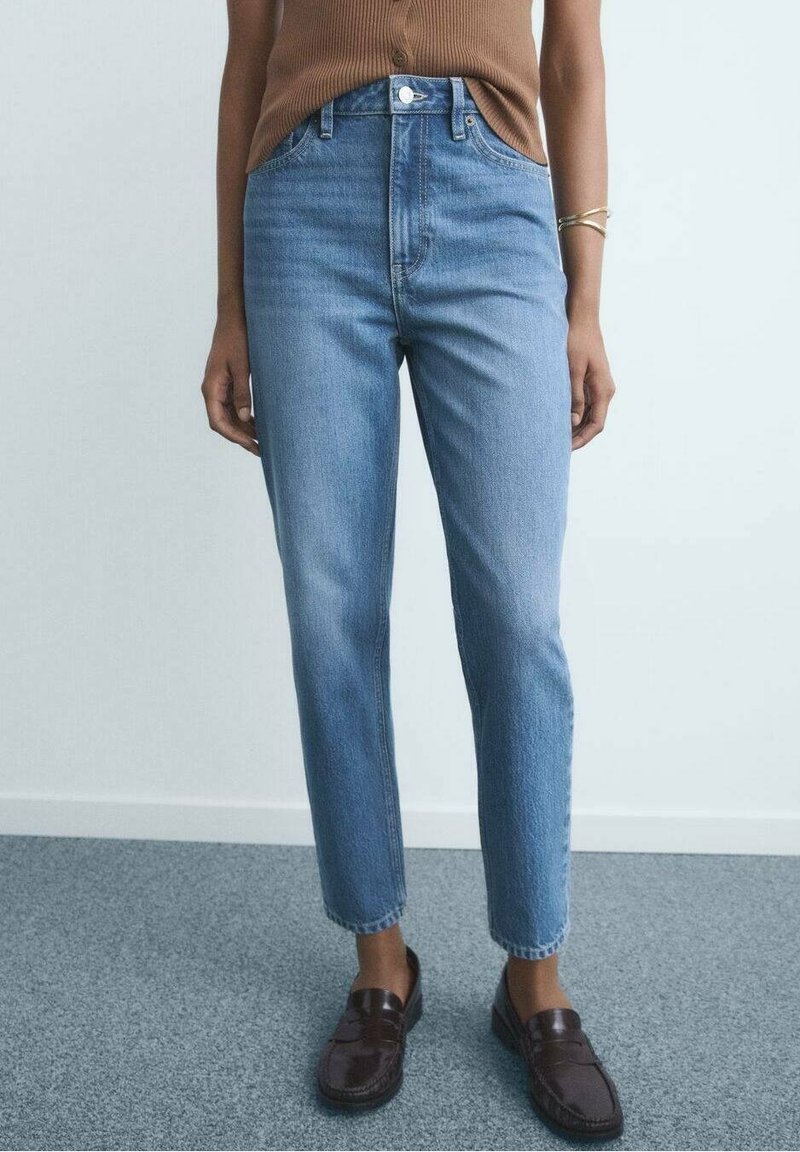 Personne portant un jean bleu clair taille haute, un haut sans manches marron avec boutons, des mocassins en cuir marron et un bracelet en or au poignet gauche.