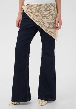 Person trägt dunkle ausgestellte Jeans, beigen floralen Spitzenschal um die Taille gebunden und beige gewebte Schuhe mit Absatz vor einfarbigem Hintergrund.