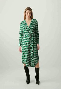 Vestido verde envolvente com padrões triangulares em preto e branco, mangas longas e cintura ajustada. Estilizado com botas pretas até ao joelho.