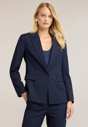 Lyshåret kvinde iført en mørkeblå skræddersyet blazer og bukser med gyldne statements øreringe, stående med den ene hånd i lommen mod en ensfarvet baggrund.