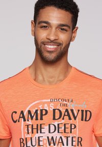 Camiseta de algodón naranja con un estampado gráfico que incluye el texto "DESCUBRE LAS ISLAS CAMP DAVID EL AGUA PROFUNDA" en negro y azul claro.