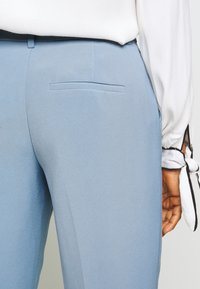 Närbild av en person som bär ljusblå skräddarsydda byxor och en vit långärmad blus med svarta detaljer och knytning vid  ärmarna.