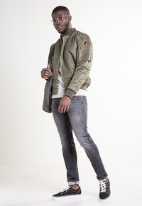 Giacca bomber verde oliva con polsini a coste, felpa grigia chiara sotto, jeans scuri e sneakers nere con lacci bianchi.