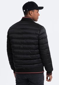 Giubbotto nero imbottito con design a trama orizzontale, polsini e colletto a coste. Strisce di accento in rosso e bianco sul collo e sull'orlo.