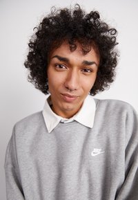 Siv hoodie z belim Nike logotipom, z teksturirano površino in kontrastnim belim ovratnikom. Vidni kodrasti lasje in prebadanje septuma.