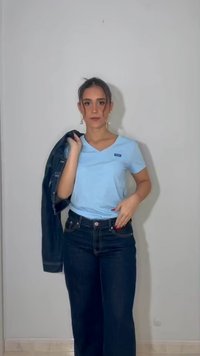 Γυναίκα με σκούρα μαλλιά που φοράει ανοιχτό γαλάζιο μπλουζάκι και σκούρο τζιν κρατάει τζιν μπουφάν στον ώμο της μπροστά σε μια απλή λευκή επιφάνεια.