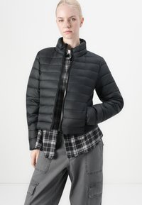 Svart puffjakke med høy krage, med horisontal quilt. Båret over en grå rutete flanellskjorte og grå cargo-bukser.