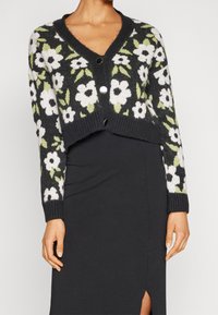 Schwarze zugeschnittene Strickjacke mit weißem und grünem Blumenmuster, V-Ausschnitt und zwei schwarzen Knöpfen. Über einem schwarzen Kleid getragen.