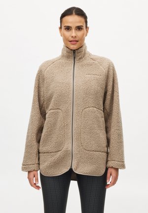 Frau trägt eine beige Sherpa-Fleecejacke mit Reißverschluss, hohem Kragen und großen Fronttaschen, kombiniert mit schwarzen strukturierten Leggings.