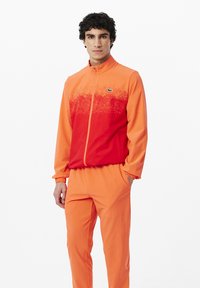 Tuta in gradiente arancione e rosso, con giacca zip-up dotata di colletto alto e polsini elasticizzati, abbinata a pantaloni coordinati.