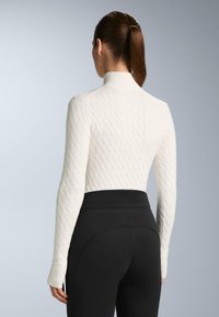 Kvinde med brun hestehale iført en tekstureret hvid rullekravetrøje og højttaljede sorte bukser, vist bagfra mod en lysegrå baggrund.