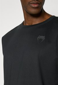 T-shirt noir à manches courtes en coton, avec un logo en relief subtil sur le côté gauche supérieur et un col rond.