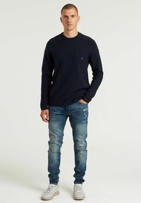 Camicia a maniche lunghe navy con tasca, abbinata a jeans chiari strappati e sneaker bianche. Design semplice con una vestibilità rilassata.