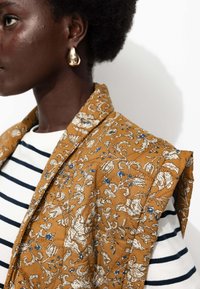 Profil d'une personne portant une boucle d'oreille en forme de goutte d'or, un gilet matelassé floral moutarde sur un haut blanc à rayures noires.