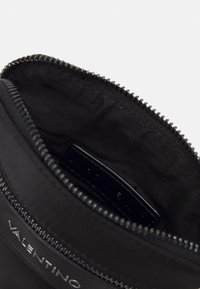 Pochette en tissu noir avec une surface texturée, fermeture éclair et une étiquette de logo visible à l'intérieur. Intérieur doublé d'un matériau lisse.