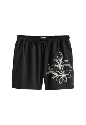 Hemdloser Mann, der schwarze Badeshorts mit Blumenstickerei trägt, steht barfuß auf einem Felsen am Meer unter klarem blauem Himmel.