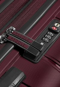 Valigia bordeaux con superficie testurizzata, dotata di cerniere metalliche, una combinazione di lucchetto e accenti di hardware neri a contrasto.