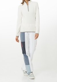 Persona vestida con un pulóver blanco de cremallera hasta el cuarto y pantalones en bloques de color en blanco, gris y azul marino, con zapatillas de colores claros.