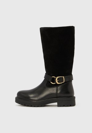 Botte noire mi-mollet avec pied en cuir, tige en daim, semelle épaisse et bride décorative à boucle dorée sur un fond clair uni.