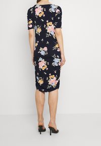 Nainen, joka on pukeutunut tiukkaan tummansiniseen mekkoon, jossa on pinkkejä, keltaisia ja sinisiä kukkakuvioita, seisoo paljain jaloin varpaillaan yksinkertaista valkoista taustaa vasten.