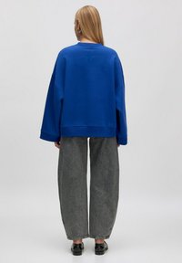 Blaues oversized Sweatshirt mit herunterhängenden Schultern, gerippten Bündchen und Saum. Kombiniert mit weiten grauen Hosen und schwarzen Schuhen.