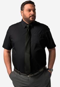 Camisa negra de manga corta con un bolsillo en el pecho izquierdo, combinada con una corbata verde oscura y pantalones grises. El portador lleva un reloj y tiene las manos en los bolsillos.