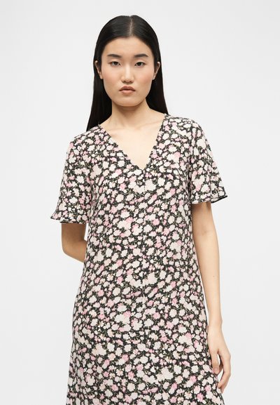 Γυναίκα με ίσια μαύρα μαλλιά που φοράει μαύρο φόρεμα με λευκό και ροζ λουλουδάτο μοτίβο, στέκεται μπροστά σε απλό λευκό φόντο.