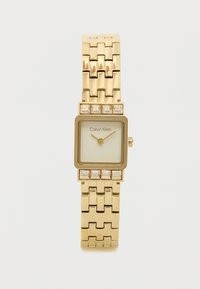 Montre-bracelet carrée en or avec un bracelet en maillons métalliques, présentant un cadran blanc et de petits accents en cristal sur la lunette. Marque : Calvin Klein.