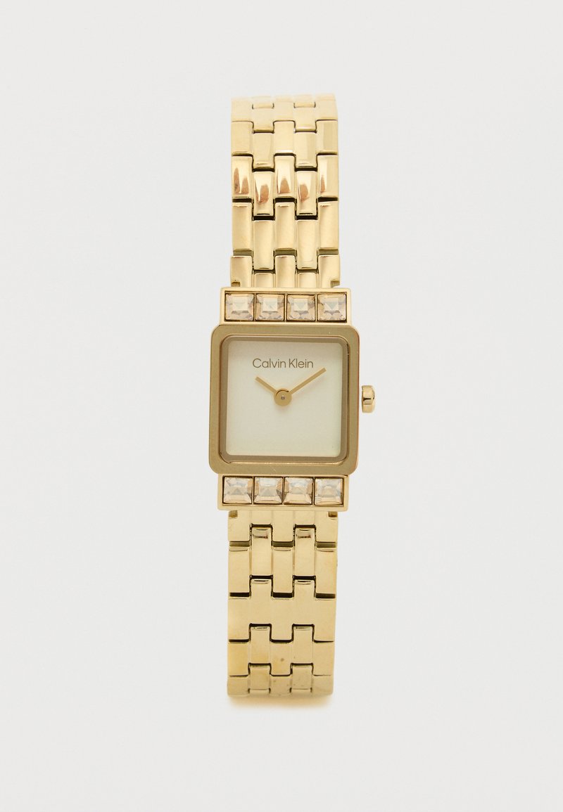 Montre-bracelet carrée en or avec un bracelet en maillons métalliques, présentant un cadran blanc et de petits accents en cristal sur la lunette. Marque : Calvin Klein.