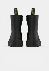 Bottines en cuir noir avec une finition texturée, semelle extérieure en caoutchouc épaisse, couture jaune et une languette portant l'inscription "avec des semelles rebondissantes".