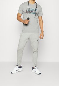 Light gray Nike t-shirt with graphic logo, paired with light gray sweatpants. White sneakers with navy accents. Hand holding a jump rope.
