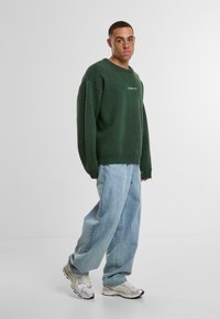 Zeleni pulover s mekanom teksturom, oversized krojem i rebrastim rukavima, uparen s svijetlosindžastim širokim trapericama i srebrnim tenisicama.