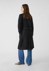 Svart ull trenchcoat med knytbälte, långa ärmar och rak silhuett, i kombination med lösa blå denimjeans och spetsiga skor.