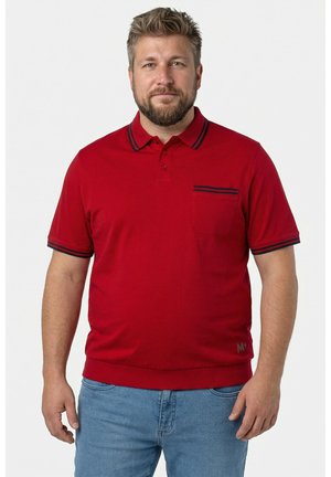 Mężczyzna z krótkimi włosami i brodą, ubrany w czerwoną koszulkę polo z czarnym wykończeniem oraz niebieskie dżinsy, stojący na jednolitym białym tle.