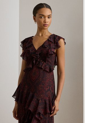 Tumejuustega naine, kellel on madal kukkel, kannab tumeda paisleemustrilise volangilise kleiti, millel on V-kaelus ja varrukateta disain, seisab halli tausta ees.