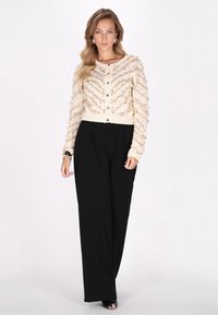 Cárdigan crema con adornos dorados, ajustado en la cintura, combinado con pantalones de pierna ancha negros. Textura suave y detalles de cierre con botones.