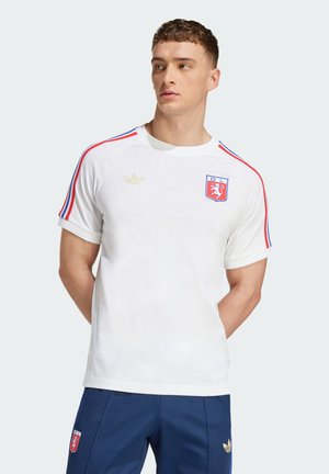 Ung mand iført en hvid sportstrøje med røde og blå striber på ærmerne og blå bukser med sportslogoer, stående med hænderne bag ryggen.