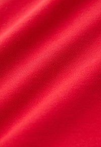 Krupan plan svijetlocrvene teksturirane tkanine s malim tkanim uzorkom i mekanim naborima koji stvaraju blage sjene.