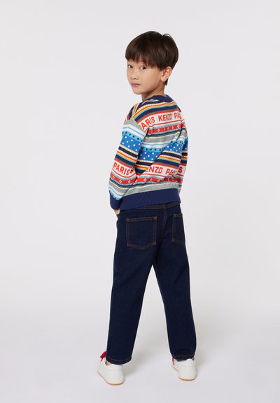 Pull en tricot avec des rayures multicolores et le texte "KENZO PARIS", associé à un jean bleu foncé et des baskets blanches avec des accents rouges.
