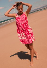 Robe sans manches rose et blanche à motif floral, en tissu léger, évasée à l'ourlet. Décor de plage avec sable et océan en arrière-plan.