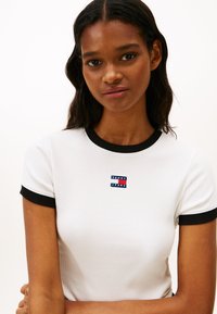 Λευκό ριπ κοντομάνικο μπλουζάκι με μαύρη επένδυση, με μικρά μπλε και κόκκινα λογότυπα στο στήθος. Κοντά μανίκια και εφαρμοστή γραμμή.